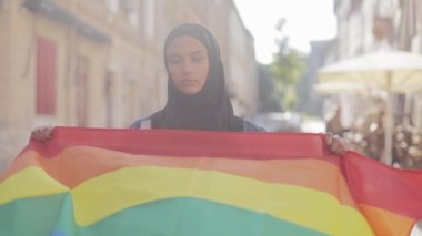 Eski sokak arka planda duran LGBT bayrağı ile başörtüsü takan güzel genç Müslüman kadın portresi. Lezbiyenlik ve İslam kavramı.
