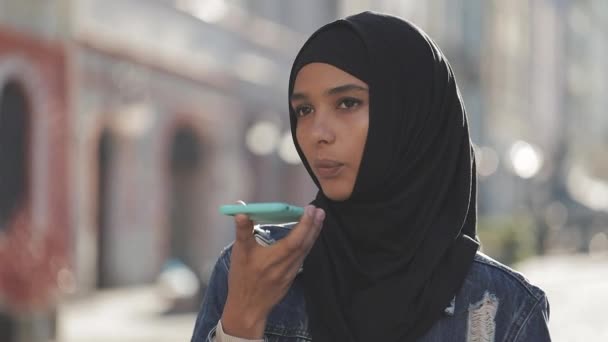 Молодая мусульманка отправляет голосовое сообщение по мобильному телефону на улице, разговаривая с мобильным ассистентом. Девушка, использующая распознавание голоса на смартфоне, диктует мысли, голосовое сообщение — стоковое видео