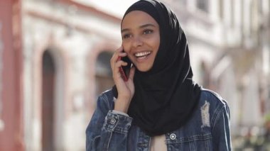 Şehirde bir smartphone arkadaşlarınızla konuşan hijab başörtüsü giyen genç Müslüman kadın, bir Bayan siyah hijab eski sokak boyunca yürür.