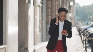 Kulaklıklı genç mutlu Afro-Amerikan iş kadını, akıllı telefondaki müziği dinleyip dans ederken şehir caddesinde yürüyor. Açık.