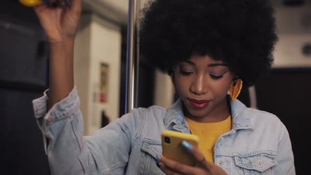 Mosolygó fiatal afro-amerikai nő rendelkezik a kapaszkodó és smartphone használata a tömegközlekedés. Éjszakai. Esőzés és városi világítás háttere. — Stock videók