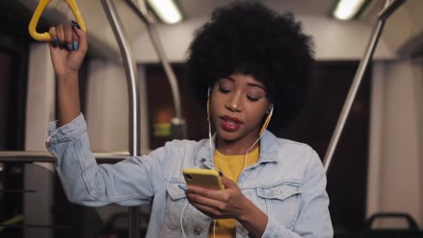 Portret młodej afroamerykańskiej kobiety noszących i ze słuchawkami słuchających muzyki, śpiewających i zabawnych tańców w transporcie publicznym. Trzyma poręcz. — Wideo stockowe