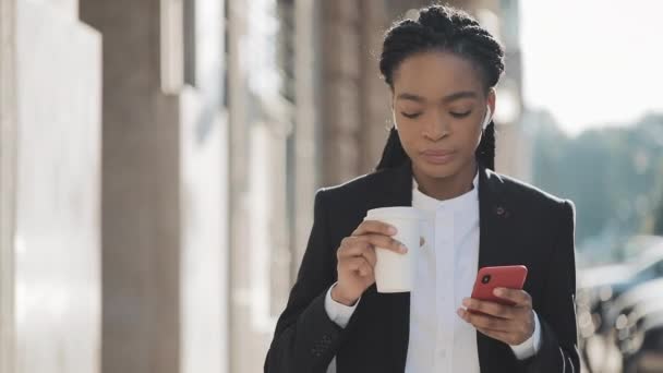 Retrato de una joven mujer de negocios afroamericana en traje, caminando por la ciudad, tomando café y usando un smartphone. Concepto: nuevo negocio, comunicación, jornada laboral, freelancer . — Vídeo de stock