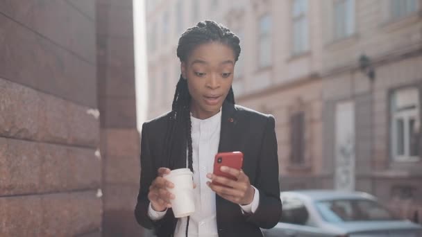 Portrét mladé africké americké obchodní ženy v obleku, který stojí na ulici, pije kávu a používá smartphone. Ženská profesionálka v obleku s dobrými zprávami o smartphone. — Stock video