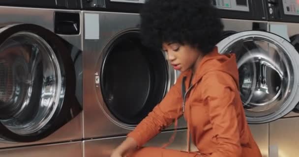 Une jeune Afro-Américaine s'assoit devant une machine à laver et charge la laveuse de linge sale. Laverie publique en libre-service . — Video