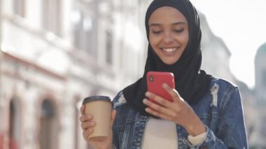 , Akıllı telefon ve içme kahvesi kullanarak, şehir merkezinde yürüyüş hijab başörtüsü giymiş Müslüman kadın smilng. İletişim, online alışveriş, sosyal ağ konsepti.