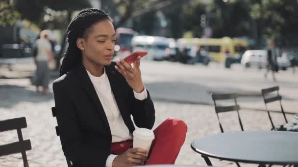 Mulher de negócios afro elegante enviando mensagem de voz de áudio no celular ao ar livre falando com assistente móvel. Senhora de negócios sentada no terraço de verão no café acolhedor. Ao ar livre . — Vídeo de Stock