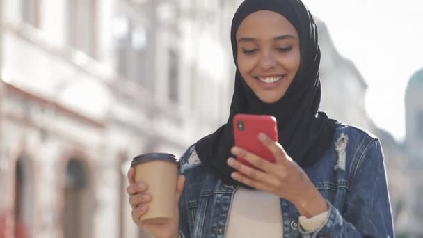 Mulher muçulmana sorrindo usando lenço de cabeça hijab andando no centro da cidade, usando smartphone e bebendo café. Comunicação, compras online, conceito de rede social . — Vídeo de Stock