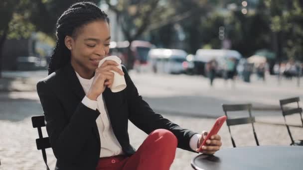 아늑한 카페의 여름 테라스에 앉아 커피를 마시는 스마트 폰을 사용하는 세련된 아프로 비즈니스 여성. 비즈니스 레이디, 모바일 기술을 즐기는 소셜 미디어에 메시지를 공유. — 비디오