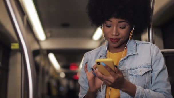 Retrato de mujeres afroamericanas jóvenes en auriculares escuchando música y navegando en el teléfono móvil en el transporte público. Fondo de luces de ciudad. Movimiento lento . — Vídeo de stock