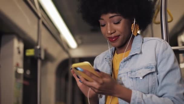 Retrato de jovens afro-americanas em fones de ouvido ouvindo música e navegando no celular em transportes públicos. Luzes da cidade fundo. Movimento lento . — Vídeo de Stock