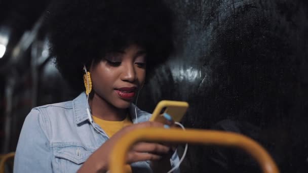 Jovem mulher afro-americana atraente usando smartphone equitação no transporte público. Boa noite. Cidade luzes fundo . — Vídeo de Stock