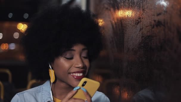 公共交通機関でスマートフォンを使用して、窓の外を見て魅力的な若いアフリカ系アメリカ人の女性。夜の時間。都市ライトの背景. — ストック動画