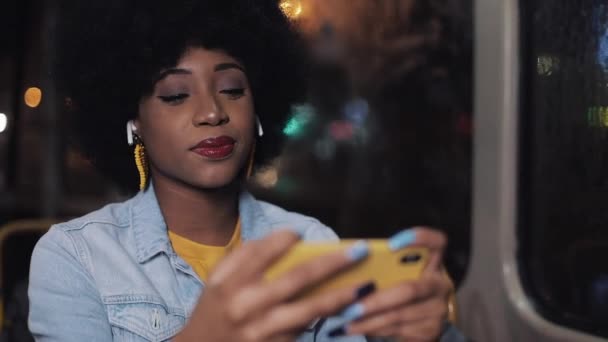 年轻的非洲裔美国妇女观看视频在智能手机骑在公共交通。夜晚时间。特写。城市灯光背景. — 图库视频影像