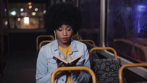 Mujer afroamericana feliz montando en el transporte público, cierra el libro después de leer y mirar por la ventana. Chica escucha música. Ciudad luces fondo . — Vídeo de stock