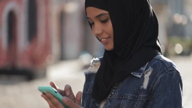 Genç gülümseyen Müslüman kadın şehir merkezinde hijab başörtüsü ayakta giyen ve akıllı telefon kullanarak. İletişim, online alışveriş, sosyal ağ konsepti.