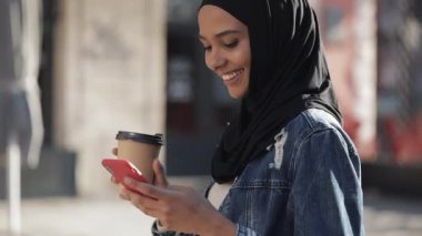 , Akıllı telefon ve içme kahvesi kullanarak, şehir merkezinde yürüyüş hijab başörtüsü giymiş Müslüman kadın smilng. İletişim, online alışveriş, sosyal ağ konsepti.