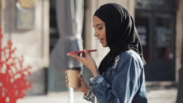 若いイスラム教徒の女性は、モバイルアシスタントに話す屋外で携帯電話で音声メッセージを送信します。スマートフォンの音声認識を使用して女の子は、思考、音声ダイヤルメッセージを指示します — ストック動画