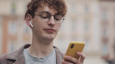 Mutlu gözlüklü hipster adam sosyal ağlarda arkadaşları ile sohbet ve müzik dinlemek, şehir sokakta yürürken modern akıllı telefon cihazı nı kullanarak. Kentsel yaşam tarzı.