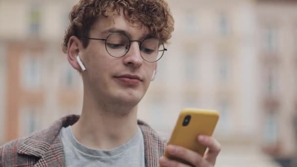 Joyeux hipster souriant avec des lunettes bavarder avec des amis sur les réseaux sociaux et écouter de la musique, en utilisant un smartphone moderne tout en marchant dans la rue de la ville. Mode de vie urbain . — Video