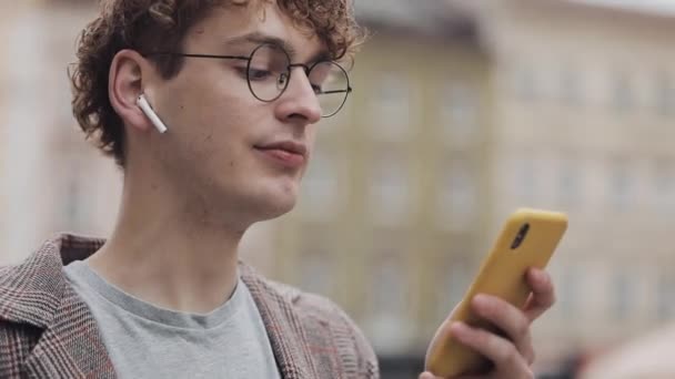 Joyeux hipster souriant avec des lunettes bavarder avec des amis sur les réseaux sociaux et écouter de la musique, en utilisant un smartphone moderne tout en se tenant dans la rue de la ville. Mode de vie urbain . — Video
