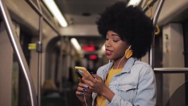 Retrato de mujeres afroamericanas jóvenes en auriculares escuchando música y navegando en el teléfono móvil en el transporte público. Fondo de luces de ciudad. Movimiento lento . — Vídeo de stock