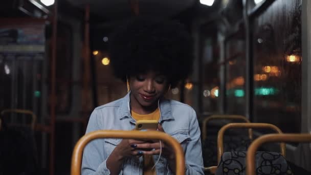 Jovem mulher afro-americana atraente usando smartphone montando em transportes públicos e olhando pela janela. Boa noite. Cidade luzes fundo . — Vídeo de Stock