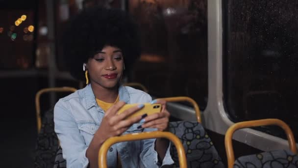 Giovane donna afroamericana che guarda video sullo smartphone cavalcando nei trasporti pubblici. Di notte. Primo piano. Luci della città sfondo . — Video Stock