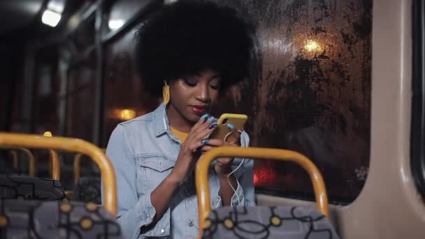 Atractiva mujer afroamericana joven utilizando smartphone a caballo en el transporte público. Noche. Ciudad luces fondo . — Vídeos de Stock