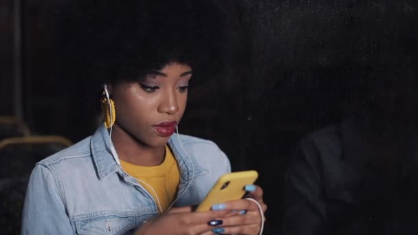 Attraktive junge afrikanisch-amerikanische Frau, die mit dem Smartphone in öffentlichen Verkehrsmitteln unterwegs ist. Nachts. Hintergrund Stadtbeleuchtung. — Stockvideo