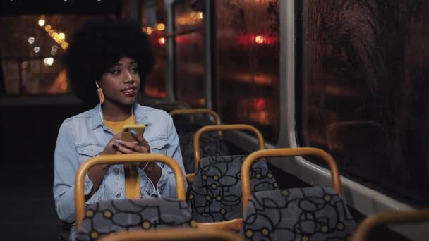 Çekici genç Afrikalı Amerikalı kadın kullanarak akıllı telefon sürme toplu taşıma ve pencereden dışarı bakıyor. Gece vakti. Şehir ışıkları arka plan. — Stok video