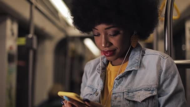 Retrato de mujeres afroamericanas jóvenes en auriculares escuchando música y navegando en el teléfono móvil en el transporte público. Fondo de luces de ciudad. Movimiento lento . — Vídeo de stock