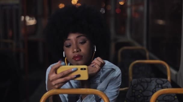 Молода афроамериканець жінка дивиться відео на смартфон верхи на громадському транспорті. Нічний час. Закри. Місто вогні фону. — стокове відео