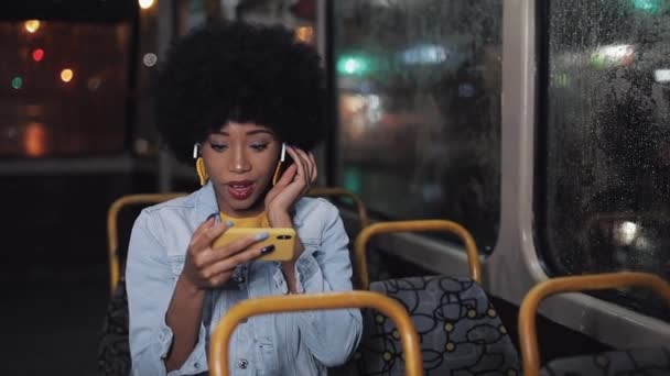 Χαμογελαστή νεαρή αφρικανική Αμερικανίδα βλέποντας αστεία βίντεο στο smartphone ιππασία στις δημόσιες συγκοινωνίες. Νύχτα. Γκρο πλαν. Φόντο φώτα πόλης. — Αρχείο Βίντεο