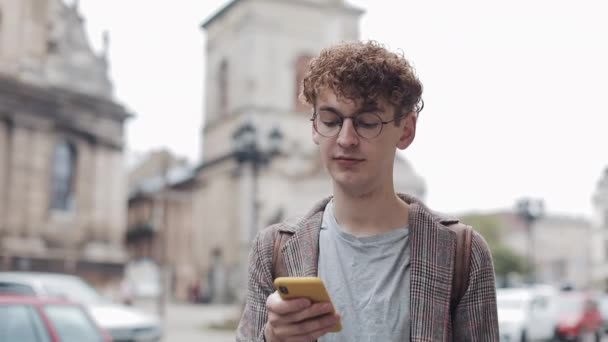 Glücklich lächelnder Hipstertyp mit Brille, der mit Freunden in sozialen Netzwerken plaudert und moderne Smartphone-Geräte benutzt, während er auf der Straße in der Stadt spaziert. Kommunikation, Student, Tourismuskonzept. urbaner Lebensstil. — Stockvideo