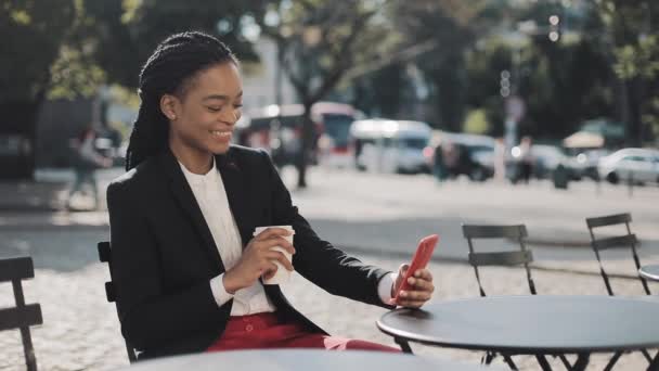 Stylish afro business woman using smartphone drinking coffee sitting on the summer terrace in cozy cafe. Business lady,sharing messages on social media enjoying mobile technology. — Stock Video