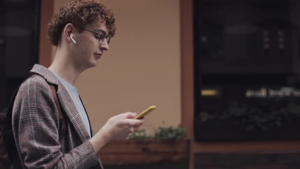 Happy smiling hipster guy with glasses chatting with friends at social networks and listen music, using modern smartphone device while standing on the city street. Urban lifestyle. — Stock Video