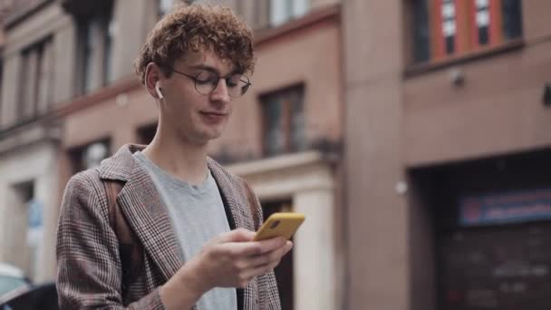 Feliz chico hipster sonriente con gafas charlando con amigos en las redes sociales y escuchar música, utilizando un dispositivo de teléfono inteligente moderno mientras está de pie en la calle de la ciudad. Estilo de vida urbano . — Vídeo de stock