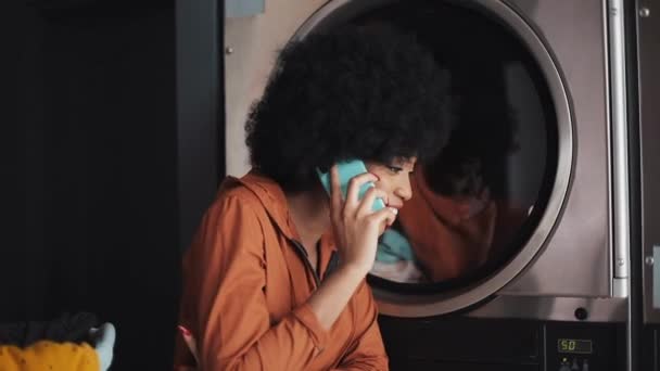 Smiling young woman talking on mobile phone at laundromat. — Stock Video