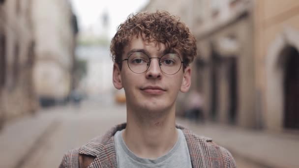 Retrato de un joven hipster con gafas de pie en la calle de la ciudad y mirando a la cámara. Turismo, viajes, concepto de estudiante. Estilo de vida urbano . — Vídeo de stock