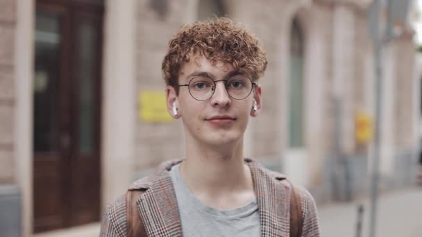 Retrato del joven hipster con auriculares inalámbricos de pie en la calle de la ciudad y mirando a la cámara. Turismo, viajes, concepto de estudiante. Estilo de vida urbano . — Vídeos de Stock