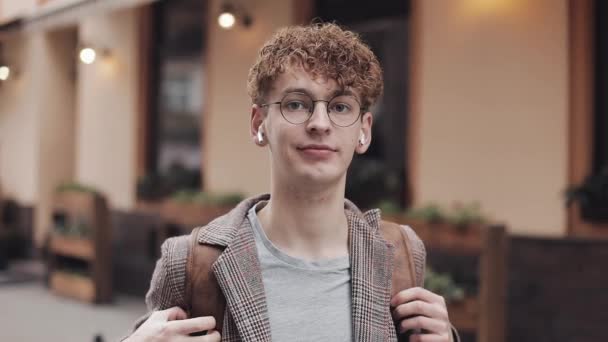 Portrét mladého hipstera, který má bezdrátové sluchátka na ulici a dívá se do kamery. Turistická, cestovní, Studentská koncepce. Městský životní styl. — Stock video