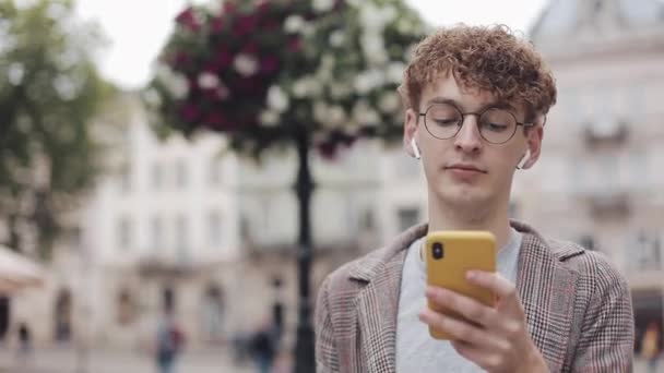 Jovem hipster com óculos conversando com amigos em redes sociais e ouvir música, usando o dispositivo de smartphone moderno enquanto caminha na rua da cidade. Estilo de vida urbano . — Vídeo de Stock