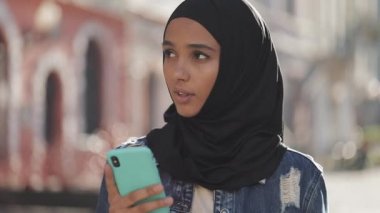 Başörtüsü takan genç müslüman kadın şehir merkezinde dikiliyor ve akıllı telefon kullanıyor. İletişim, online alışveriş, sosyal ağ kavramı.