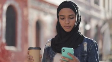 Başörtüsü takan genç müslüman kadın şehir merkezinde yürüyerek, akıllı telefon kullanarak ve kahve içiyor. İletişim, online alışveriş, sosyal ağ kavramı.