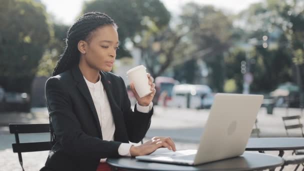 Glimlachend Afro-Amerikaanse zakenvrouw zittend in café op het zomerterras, koffie drinken en aan de laptop werken. Zakelijk, werkend, freelance concept. — Stockvideo