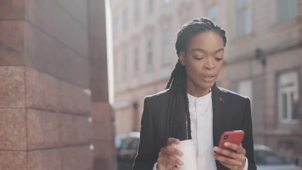 Портрет молодой афроамериканской бизнесвумен в костюме, гуляющей по городу, пьющей кофе и пользующейся смартфоном. Женщина-профессионал в костюме, имеющая хорошие новости на смартфоне . — стоковое видео