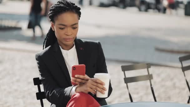 Mulher de negócios afro elegante usando smartphone beber café sentado no terraço de verão no café acolhedor. Comunicação, trabalho, conceito de rede social . — Vídeo de Stock