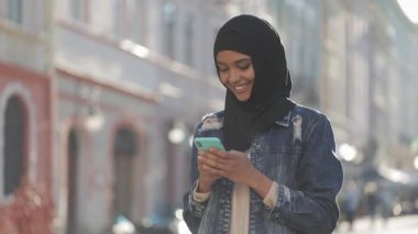 Genç gülümseyen Müslüman kadın şehir merkezinde hijab başörtüsü ayakta giyen ve akıllı telefon kullanarak. İletişim, online alışveriş, sosyal ağ konsepti.