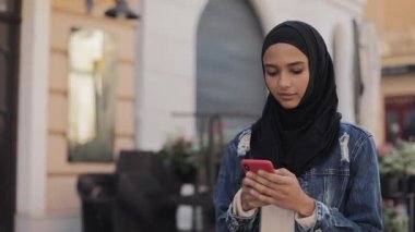 Başörtüsü takan genç müslüman kadın şehir merkezinde yürüyerek ve akıllı telefon kullanıyor. İletişim, online alışveriş, sosyal ağ kavramı.
